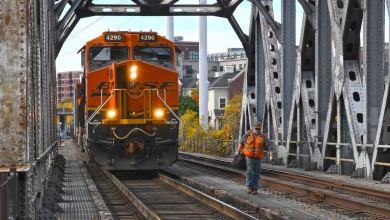 A photograph of a freight train passing through a railroad bridge. A railroad worker is standing next to the train.