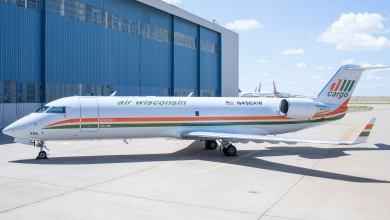 A small Air Wisconsin cargo jet sits on tarmac next to big hangar on bright day.