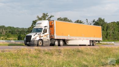 A white Schneider daycab pulling an orange Schneider dryvan trailer on a highway