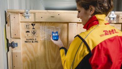 A woman wearing a gold-and-red DHL jacket attaches a device to a wood shipping crate.