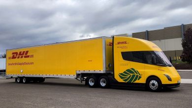 A yellow DHL Supply Chain Tesla Semi electric truck, branded with the DHL logo and a green leaf emblem for sustainability, parked in front of a modern warehouse under overcast skies in California.