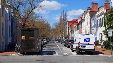 A brown UPS van on the left side of a city neighborhood street, with a USPS van on the right side on a sunny day.
