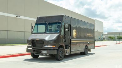 A brown UPS van is on a street next to a large side of a warehouse.