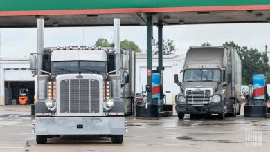 Two semi-trucks refueling at a truck stop diesel station under a canopy, illustrating the impact of rising fuel costs on carriers.