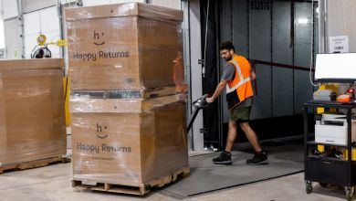 A man wearing an orange vest loads cardboard boxes labeled Happy Returns onto a truck with a pallet moving tool.