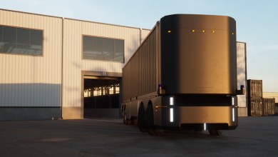 abless Humble Hauler autonomous electric truck pulling a shipping container outside a large industrial warehouse loading dock at sunrise