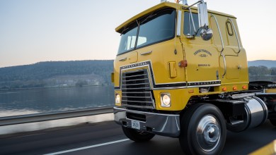 Classic bright yellow International Transtar II semi-truck cab with Truck Parking Club and Ringgold, GA branding, driving on a scenic highway beside a calm lake with rolling green hills in the background.