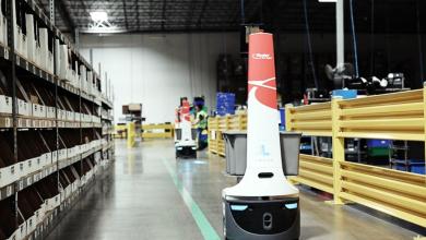 Ryder Locus autonomous mobile robot with red-and-white branding and attached bins navigating a modern warehouse aisle lined with tall industrial shelving, yellow safety barriers, and green floor markings, while another Locus robot and a worker operate in the background.