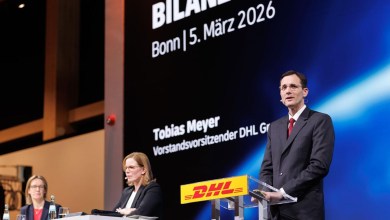 An executive speaks at a podium with DHL sign on it and a slide presentation on a big screen behind him.