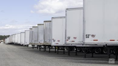 Long row of white semi-trailers parked on landing gear in a large gravel lot under a partly cloudy sky.