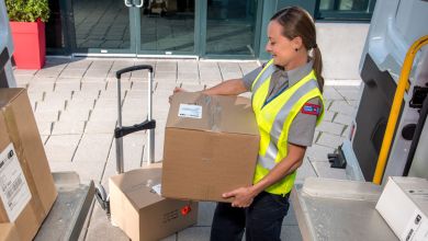 A female Canada Post letter carrier lifts a box to hand truck during a delivery stop.