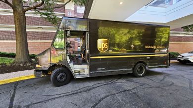 A brown UPS delivery van is parked under the canopy of an apartment building while the driver delivers packages inside.