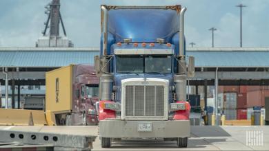 Blue semi-truck facing forward at a busy freight terminal with yellow shipping container, port cranes, and other trucks visible in the background