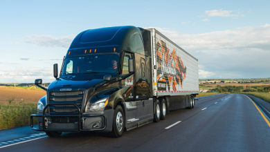 Black Hirschbach Freightliner semi-truck with branded trailer driving on a rural highway under a partly cloudy sky