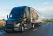 Black Hirschbach Freightliner semi-truck with branded trailer driving on a rural highway under a partly cloudy sky